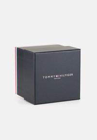 Boîte carrée noire avec le texte blanc "TOMMY HILFIGER WATCHES" sur le devant, avec une rayure d'accent rouge et bleue sur le bord supérieur.