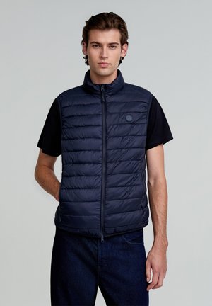 Marineblauwe gewatteerde vest met een hoge kraag, verticale stiksels en een ritsluiting. Bevat een logo-detail op de borst. gedragen over een zwart T-shirt.