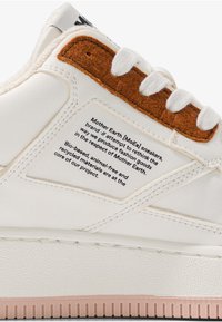 Witte sneaker met een gestructureerd oppervlak, bruine suède accenten op de tong en een label met details over milieuvriendelijke materialen en productie.
