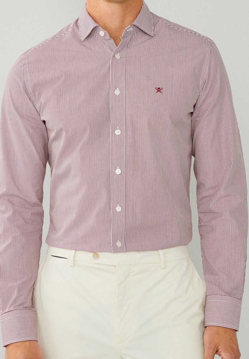 Chemise à boutons rayée rouge et blanche avec un col classique et un emblème brodé ; tissu léger ; manches longues ; portée avec un pantalon beige.