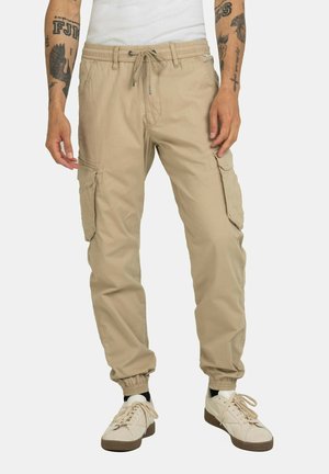 Pantalones cargo beige con puños elásticos, cintura con cordón y múltiples bolsillos laterales. Combinados con zapatillas blancas y calcetines negros hasta el tobillo.