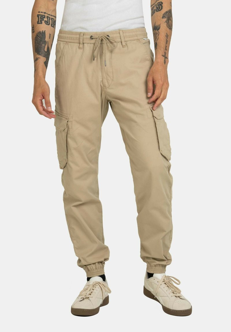 Beige Cargo-Hosen mit elastischen Bündchen, Kordelzugbund und mehreren Seitentaschen. Getragen mit weißen Turnschuhen und schwarzen Knöchelstrümpfen.