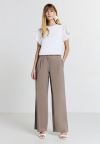 Top bianco con maniche a volant in pizzo traforato, abbinato a pantaloni beige a vita alta e fit morbido. Il modello indossa sandali bianchi con apertura davanti.