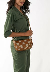 Sac bandoulière marron avec un motif à carreaux, chaîne dorée, forme rectangulaire et texture lisse. Porté sur une combinaison verte.
