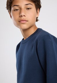 Laivastonsininen neulepaita pyöreällä kauluksella ja teksturoidulla kankaalla, istuva malli ja resorikuvioidut hihansuut. Ei näkyviä kuvioita tai koristeita.