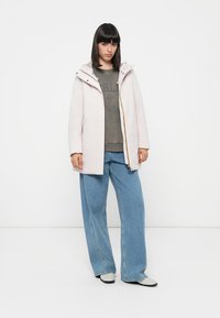 Parka de color rosa claro, acolchada y con capucha, que cuenta con un cierre frontal con un detalle dorado. Lleva puesta una sudadera gris y unos vaqueros anchos azules.