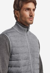 Szara pikowana kamizelka z wysokim prążkowanym kołnierzem i zapięciem na zamek, z kontrastującymi szarymi rękawami i gładką teksturą. Lekka konstrukcja.