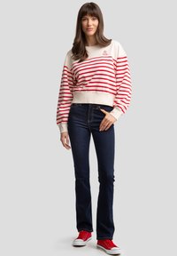 Sudadera blanca y roja a rayas con mangas anchas, combinada con jeans acampanados azul oscuro y zapatillas rojas, sobre un fondo gris.