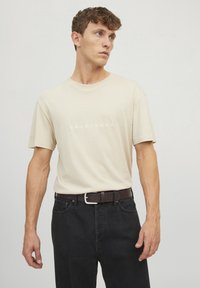 Camiseta de algodón beige con cuello redondo y mangas cortas, con la palabra "ORIGINALS" impresa en texto blanco en la parte frontal, combinada con jeans de mezclilla oscuros y un cinturón de cuero marrón.