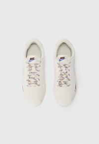 Balti ādas kedas ar daudzkrāsainām lentēm, noapaļota purngala forma, mīksts apkakles daļa un diskrēts Nike zīmols uz mēlītes un sāniem.