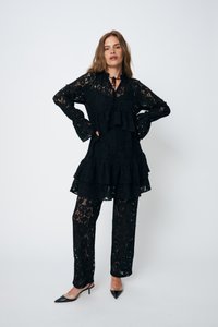 Abito in pizzo nero con strati arricciati, maniche lunghe e dettaglio con fiocco, abbinato a pantaloni in pizzo coordinati. Presenta motivi floreali trasparenti.