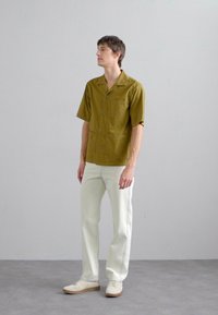 Kurzarm-Hemd in Olivgrün mit zwei aufgesetzten Vordertaschen, kombiniert mit weißen Hosen und beigen Sneakers, vor einem neutralen Hintergrund stehend.