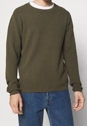 Man die een olijfgroene, gestructureerde trui met lange mouwen draagt over een wit T-shirt en een blauwe spijkerbroek, staand tegen een effen achtergrond.