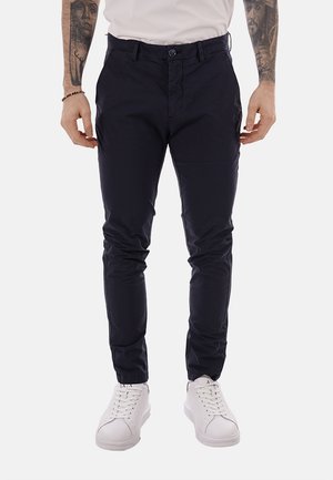 Uomo che indossa pantaloni aderenti blu navy, sneaker bianche e una camicia bianca, con braccia tatuate e un braccialetto di perline visibile.