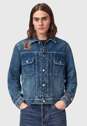 Jonge persoon met schouderlang bruin haar, gekleed in een blauwe spijkerjasje met patches van vogels en letters, handen in de zakken.