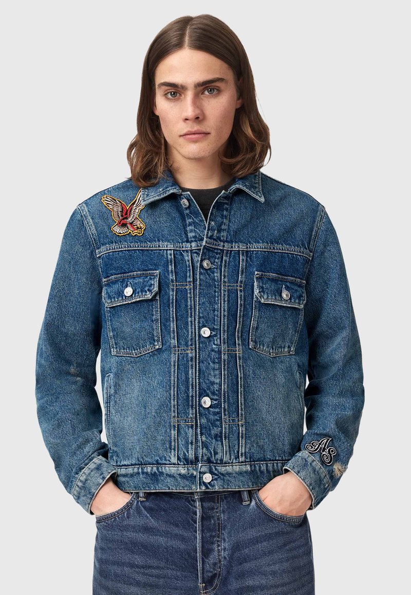 Ung person med skulderlangt brunt hår iført en blå denimjakke med fugle- og bogstavsikker, hænderne i lommerne.