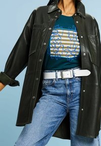 Svart läderjacka med tryckknappar, bärs över en teal-färgad skjorta med blommönster och beige ränder, kombinerad med ljusblå denim och ett vitt bälte.