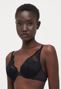 Bralette in pizzo nero con ricami floreali, pannelli trasparenti, spalline regolabili e una texture liscia con rifiniture dettagliate.