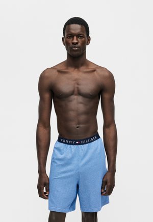 Hombre sin camisa de pie mirando hacia adelante, lleva pantalones cortos Tommy Hilfiger azul claro con cinturilla azul marino y logo sobre un fondo liso.