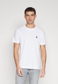 T-shirt bianco in cotone con collo rotondo e maniche corte, con un piccolo emblema blu navale sul lato sinistro del petto. Pantaloni grigi indossati in basso.