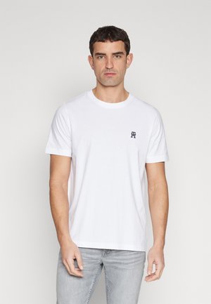 T-shirt bianco in cotone con collo rotondo e maniche corte, con un piccolo emblema blu navale sul lato sinistro del petto. Pantaloni grigi indossati in basso.