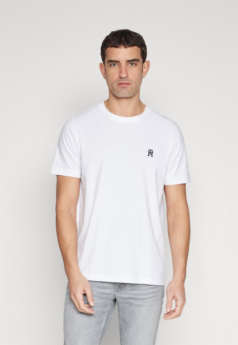 T-shirt bianco in cotone con collo rotondo e maniche corte, con un piccolo emblema blu navale sul lato sinistro del petto. Pantaloni grigi indossati in basso.