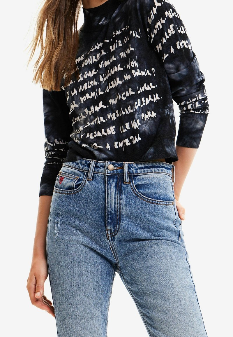 Femme portant un chemisier noir à manches longues avec des motifs de texte manuscrit blanc et un jean taille haute bleu clair.