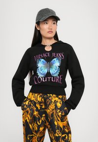 Melnā saspiestā sporta krekla ar tauriņu grafiku un tekstu "Versace Jeans Couture". Saskaņots ar melnām un zeltainām rakstainām biksēm. Nēsāta pelēka cepure.