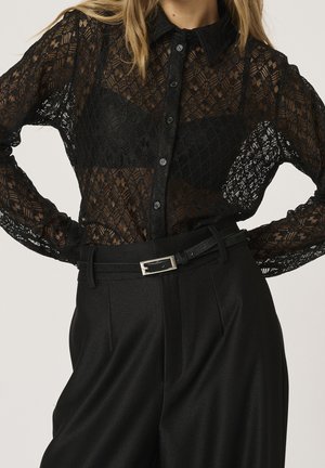 Kobieta ubrana w czarną koronkową bluzkę na guziki, pod którą ma dopasowany czarny bralet, oraz czarne spodnie z wysokim stanem i paskiem.