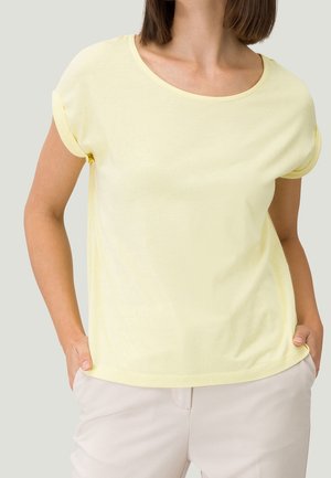Femme portant un t-shirt jaune clair uni à manches courtes retroussées et un pantalon beige, mains dans les poches, fond neutre.