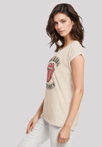 Beige Baumwoll-T-Shirt mit aufgerollten Ärmeln, vorne mit großem, aufgedrucktem Rolling-Stones-Logo mit roten Lippen und schwarzem Text.