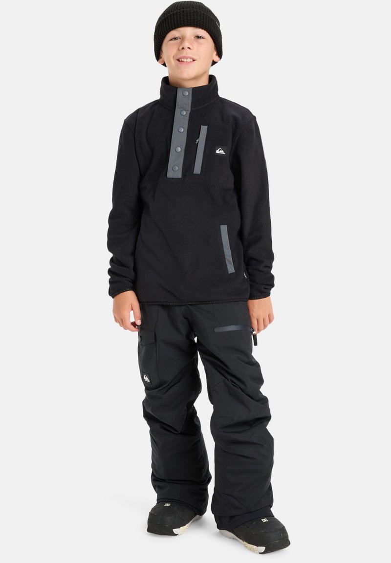 Schwarzer Fleece-Pullover mit grauen Knöpfen und Taschen, kombiniert mit schwarzen Schneehosen mit grauen Akzenten, getragen mit einer schwarzen Mütze.
