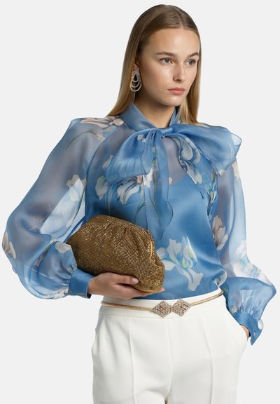 Sheer blue blouse with floral pattern, oversized bow at neck, balloon sleeves. Gold metallic clutch, white trousers with jeweled belt detail.