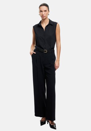 Musta ilmanhiha blouse, jossa on kaulus, napitettava etuosa, yhdistetty korkeavyötäröisiin mustiin housuihin ja teräväkärkisiin mustiin korkeakorkoisiin kenkiin.