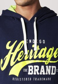 Mann trägt einen navyblauen Hoodie mit weißen Kordeln und einem leuchtend gelben und weißen Schriftzug "Indigo Heritage Brand Registered Trademark."