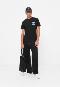 Schwarzes Baumwoll-T-Shirt mit einem grafischen Design auf der linken Brust, kombiniert mit schwarzen Jeans und einer grauen Kappe, und trägt eine schwarze Tragetasche.