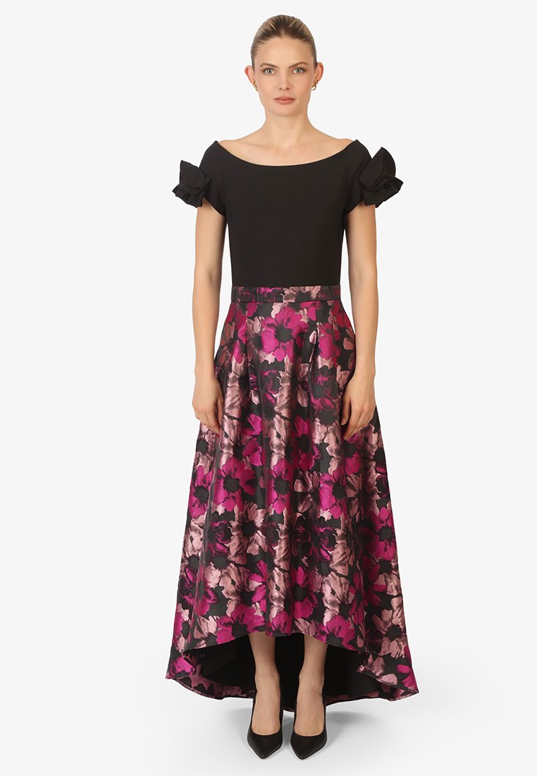 Haut noir décolleté avec des manches volantes, associé à une jupe à imprimé floral en rose et noir, plus longue à l'arrière. Des chaussures à talons noires complètent la tenue.