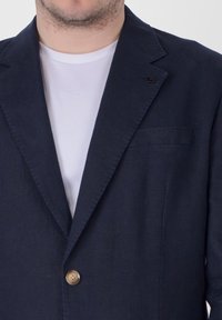 Blazer in lino blu con un solo bottone, rever a punta e taschino sul petto. Indossato sopra una t-shirt bianca, che mette in evidenza un tessuto testurizzato.