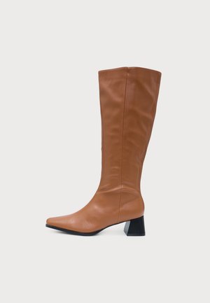 Botte en cuir marron montant jusqu’au genou, à bout pointu, avec une couture latérale et un talon bloc noir, sur fond uni.