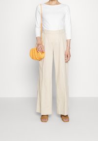 Kvinna klädd i en vit långärmad topp, beiga vida byxor, bruna vävda klacksandaler och håller en liten orange stickad crossbody-väska.