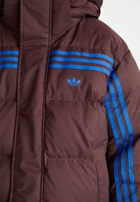 Smeđi puffer jakna s plavim horizontalnim prugama i Adidas logotipom na prsima. Materijal je gladak s kviltanom teksturom.