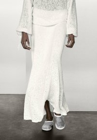 Abito bianco a maglia con maniche lunghe a campana e orlo svasato, abbinato a scarpe slip-on grigio chiaro. Il tessuto ha un aspetto texturizzato.