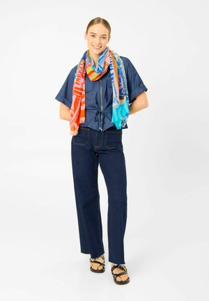 Lächelnde Frau steht und trägt eine dunkelblaue kurzärmelige Jacke, Jeans mit weitem Bein, schwarze Sandalen und einen bunten mehrfarbigen Schal um den Hals gelegt.