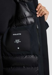 Zwarte geïsoleerde vest met een gladde, gewatteerde binnenkant, een stoffen zak met de merknaam "KHUJOTM" en een zijzak met rits.