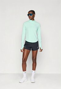 Mintgroen langärmlig topje met ritssluiting, zwarte korte broek, witte sokken, zonnebril; atletische, lichte materialen, met Nike-logo's.
