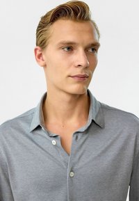 Grijze longsleeve shirt met een lichtgrijze kraag, voorzien van twee knopen en een gladde textuur. De stof heeft een subtiel patroon.