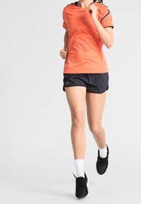 Světlé oranžové sportovní tričko s černými akcenty, v kombinaci s námořnickými šortkami. Oblečení zahrnuje bílé ponožky a černé běžecké boty.