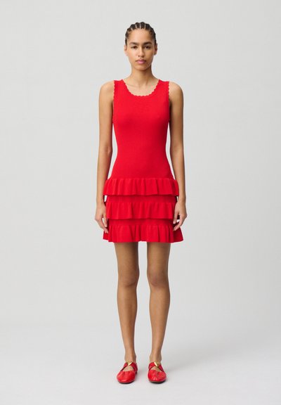 Vestito a maglia rossa a coste con scollatura ondulata, caratterizzato da tre volants sovrapposti all'orlo. La modella indossa delle scarpe rosse basse. Sfondo neutro.