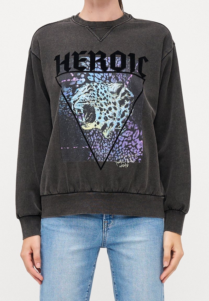 Personne portant un sweat-shirt gris foncé avec un graphisme de léopard rugissant, un cadre triangulaire et le mot "HEROIC" au-dessus, associé à un jean bleu clair.