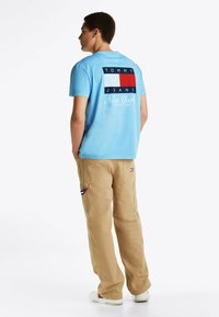 Camiseta de algodón de color azul claro con un gran logotipo gráfico en rojo, blanco y azul marino en la parte posterior. Combinada con pantalones cargo beiges.